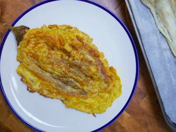 Tortilla de berenjena dorada y jugosa, servida en plato blanco con borde azul. Al lado, una bandeja con papel de horno y más berenjena.