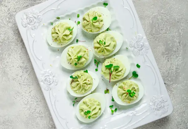 Deliciosos huevos rellenos de aguacate, decorados con brotes verdes y especias, servidos en una bandeja blanca con relieve floral.