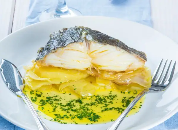 Merluza al horno con patatas, jugosa y dorada, servida sobre una cama de patatas y cebolla caramelizada, bañada en aceite de oliva y perejil picado.