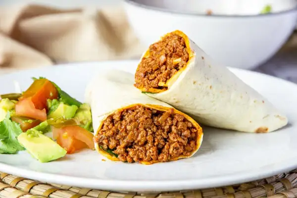 Dos burritos de carne picada, cortados por la mitad, servidos con una ensalada fresca de aguacate y tomate.