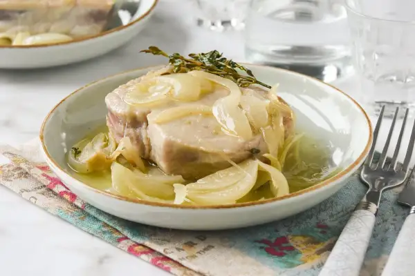 Un trozo de atún en escabeche con cebolla y tomillo, servido en un plato blanco con un tenedor al lado.