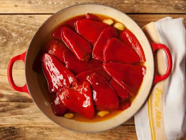 Pimientos del piquillo asados en una cazuela roja, con ajos y un paño de cocina.