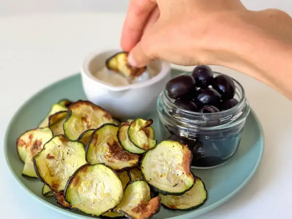 Crujientes rodajas de calabacín en airfryer, servidas con aceitunas negras y una salsa para mojar.