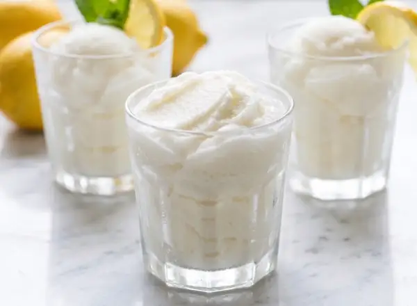 Tres vasos de sorbete de limón, refrescante y cremoso, con limones amarillos y hojas de menta al fondo.