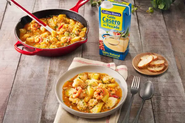 Delicioso rape a la marinera con patatas y gambas, servido en plato y sartén, junto a caldo de pescado y pan.