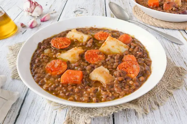 Un plato humeante de lentejas con chorizo, patatas y zanahoria, servido en un cuenco blanco sobre una mesa de madera.