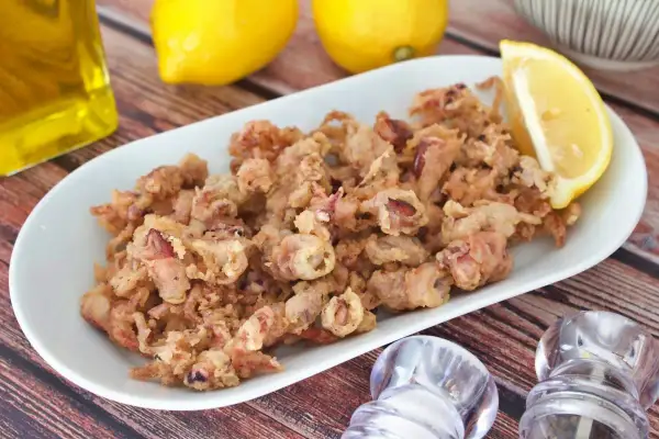 Crujientes puntillas de calamar fritas en un plato blanco, acompañadas de limón, aceite y saleros.