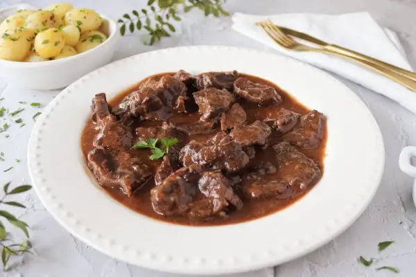 Delicioso morcillo de ternera estofado en salsa, servido en plato blanco con patatas cocidas y perejil picado.