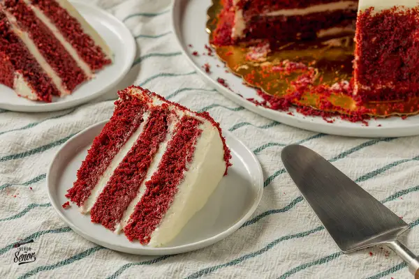 Porción de pastel red velvet con capas de bizcocho rojo y crema blanca. Otro trozo y el pastel entero en el fondo.