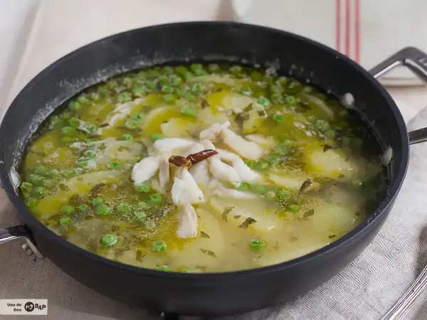 Patatas en salsa verde con trozos de pescado blanco y guisantes, servido en una sartén negra.
